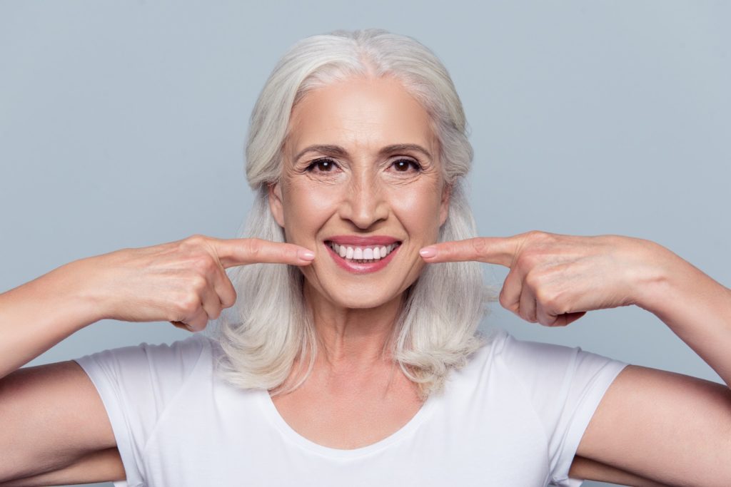 An older woman points to her bright, white smile with both hands